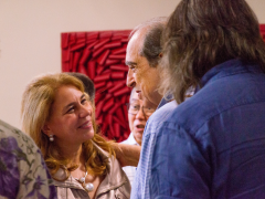 Foto de um grupo de pessoas conversando. Ao centro, uma mulher branca, de cabelos loiros e blusa bege, sorri enquanto é tocada no ombro por um homem branco, de cabelos grisalhos e camisa azul clara, que a olha e fala. À esquerda e de costas para a foto, uma mulher de blusa florida e brincos grandes. À direita, um homem de cabelos longos, vestindo camisa azul escura, também está de costas. Ao fundo, há outras pessoas e um quadro vermelho em uma parede bege.