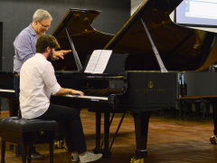 Foto de uma aula de piano. Um jovem, de cabelos castanhos e barba, está sentado de costas, com um piano preto a sua frente e tocando o instrumento. Ele veste uma camisa branca, calça preta e tenis. Ao seu lado, está um homem branco, de cabelos curtos e grisalhos, usando óculos e uma camisa azul e branca listrada. Ele observa as suas duas mãos, enquanto conversa com o jovem. O chão é de madeira e ao fundo uma parede preta e a vista parcial de um projetor