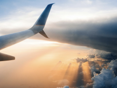 Foto da asa direita de um avião. A asa está posicionada no canto esquerdo da foto. No horizonte há nuvens contornadas por diferentes tons de laranja e sombreados de um sol poente.