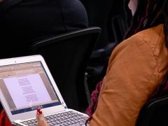 Foto de uma mulher negra lendo um texto em um laptop em seu colo. Ela tem dreads vermelhos e compridos, usa blazer caramelo e unhas vermelhas e está sentada de costas para a câmera, em um auditório. À frente dela, parte de uma cadeira e de uma pessoa de costas.