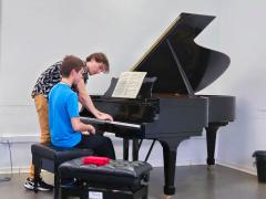 Foto de dois homens brancos jovens em frente a um piano de cauda preto. Ambos têm cabelos curtos, lisos e castanhos claros. Um deles, de camiseta azul e calça preta, está sentado em um banco preto em frente ao piano. O outro, de camiseta preta e branca e calça bege, está em pé tocando o piano. Sobre o piano, uma partitura. As paredes são brancas e, do lado deles, outro banco preto sobre o qual há um tecido vermelho.  