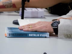 Foto de uma mão de pessoa branca repousando sobre um livro azul que está sobre uma mesa clara. O livro está com um marcador laranja e tem escrito na sua lombada “O Vazamento” e “Natalia Viana”. A mão tem uma aliança dourada no dedo anelar, um relógio preto no pulso e uma manga cinza de paletó com botões. Ao fundo e desfocada, a base de um microfone, uma caixa branca e o braço de outra pessoa branca. 