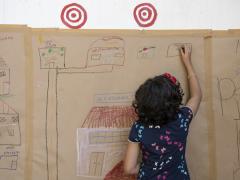 Foto de crianças e adultos participando de uma atividade artística em espaço público. As crianças estão sentadas sobre um grande papel branco, utilizando pincéis, tintas e outros materiais para pintar e desenhar. Há caixas de lápis de cor e potes de tinta ao redor. Algumas pessoas estão em pé, observando ou interagindo. O chão é de pedra.