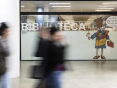 Foto da entrada da biblioteca da ECA. Sobre uma divisória de vidro, destaca-se em letras grandes e brancas a palavra ‘biblioteca’ e uma ilustração do cartunista Dorinho de uma pessoa de costas com uma mochila repleta de livros. Em primeiro plano e em desfoque, três pessoas caminham pelo saguão. 