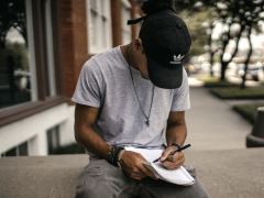 Foto de um jovem sentado em uma mureta de concreto escrevendo em um caderno apoiado no colo. Ele é negro, usa camisita cinza, calça jeans cinza, colar, pulseiras, relógio e um boné preto com logo da adidas. Ele está olhando para baixo, com a aba do boné cobrindo o rosto. O fundo da imagem está desfocado, com um prédio de tijolos vermelhos, janelas brancas e árvores na calçada.