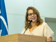 Foto de uma mulher em pé em um púlpito de madeira, fazendo um discurso. Ela é branca e tem cabelos longos, ondulados e loiros. Ela está usando óculos com armações escuras e uma blusa de cor clara. À esquerda, há uma bandeira azul. O fundo é esverdeado.