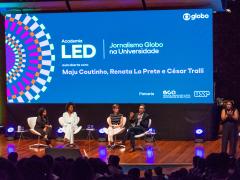 Foto de cinco pessoas em um palco diante de uma plateia. Quatro delas, duas mulheres negras, uma mulher branca e um homem, estão sentadas em poltronas brancas. O homem fala ao microfone e gesticula olhando para a plateia, enquanto as mulheres o olham. No canto direito do palco, uma mulher negra está de pé fazendo linguagem de sinais. No fundo do palco, um grande telão exibe os dizeres ‘Academia LED. Jornalismo Globo na Universidade. Aula aberta com Maju Coutinho, Renata Lo Prete e César Tralli. 