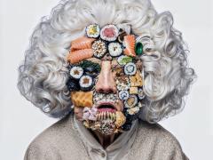 Imagem de uma mulher idosa com o rosto quase totalmente coberto por diferentes tipos de comida japonesa. Ela tem traços orientais, cabelos brancos, cacheados e volumosos na altura do pescoço e usa suéter creme e camisa branca. Do seu rosto, é possível ver apenas o nariz, os olhos e a boca. O restante está coberto por niguiris, sushis, sashimis e outros tipo de comida japonesa. O fundo é cinza claro.