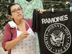 Frame de cena do filme Que horas ela volta? Uma mulher parda, de óculos retangulares, cabelos castanhos, lisos e presos, blusa vinho, avental branco com detalhes pequenos e um pano no ombro, estendendo uma camiseta preta da banda Ramones no varal. Atrás dela, há outros varais e uma parede coberta por folhas de trepadeira. 