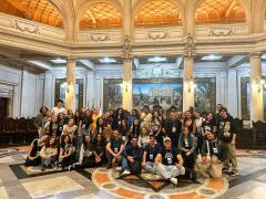 Foto de um grupo de cerca de 50 pessoas, majoritariamente mulheres jovens e brancas, todos com crachás pendurados no pescoço, dentro de um edifício antigo. O piso é feito de desenhos geométricos em mármore cinza, preto e laranja. Atrás das pessoas, colunas com luminárias e uma pintura grande pendurada na parede. O teto também é construído de abóbodas e desenhos esculpidos. 