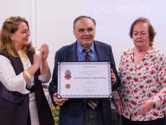 Foto de três pessoas em pé, lado a lado. No centro, um homem branco, idoso, vestindo terno escuro, camisa azul e gravata listrada, segurando e exibindo um certificado. À esquerda, uma mulher branca de cabelos loiros e longos, vestida com uma roupa preta e branca, está batendo palmas. À direita, há outra mulher branca, idosa, de cabelos castanhos curtos, vestindo uma blusa florida. Ao fundo, tela de projeção exibindo texto em azul e branco.