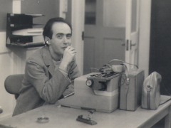 Foto em preto e branco de homem de cabelos curtos e escuros, usando paletó claro, sentado em uma cadeira atrás de uma mesa de escritório. Ele olha para a câmera enquanto leva a mão à boca. Sobre a mesa há uma máquina de escrever, uma maleta e uma bolsa de couro e um cinzeiro. Ao fundo, há portas de madeira com painéis de vidro e uma prateleira de parede com papéis. 
