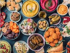 Foto a partir de cima de uma superfície azul sobre a qual estão cerca de 20 recipientes com diferentes alimentos muito coloridos: frutas secas, pastas, bolinhos fritos, pepinos, romãs, pães, entre outros. 