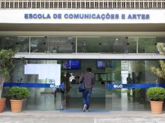 Foto da fachada de um prédio com portas de correr de vidro abertas. As portas têm uma faixa adesiva azul horizontal com o logotipo da Escola. Acima da entrada há um letreiro escrito Escola de Comunicações e Artes. A foto capta uma pessoa de costas entrando no prédio, que veste um agasalho escuro, uma calça jeans azul e está com uma bolsa pendurada no ombro esquerdo. Nas duas laterais da entrada há vasos de plantas ornamentais. No saguão interno há monitores de TV e pessoas ao fundo.