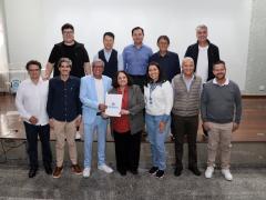 Foto de um grupo de 12 pessoas, de pé e posicionadas em duas fileiras. Sete pessoas à frente e cinco pessoas atrás, em cima de um palco de madeira, em sua maioria homens brancos. Ao centro, um homem negro, de cabelos brancos e curtos vestindo um terno azul claro e uma mulher parda, de cabelos de tamanho médio, vestindo um terno cinza e uma camisa vermelha, seguram e exibem uma pasta branca com a palavra “Suzano” impressa em azul. Ao fundo, uma tela branca e o piso de madeira.