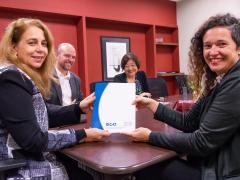 Foto de duas mulheres - ambas brancas, uma de cabelos longos e loiros e outra de cabelos castanhos cacheados - sorrindo enquanto posam segurando um documento com capa azul e branca, com os logotipos "eca" e "USP" em azul. Atrás delas, um homem branco careca e com barba e uma mulher amarela de cabelos castanhos e curtos também sorriem. Todos estão sentados em torno de uma mesa retangular de madeira. Ao fundo, uma parede vermelha com prateleiras embutidas e um quadro.
