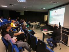 Foto de uma palestra em um auditório com cadeiras azuis, Uma plateia com cerca de 15 pessoas  assiste a uma apresentação expositiva de um homem sentado em uma mesa à  direita da foto e de costas. Ele usa fones de ouvido e boina branca, está sentado de frente para o público, apontando para uma tela grande onde um texto está projetado. Ele usa blusa preta e calça azul. O ambiente é pouco iluminado.
