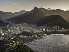 Foto da cidade do Rio de Janeiro a partir do alto. Paisagem litorânea com muitos prédios rodeados por montanhas, o mar repleto de barcos e iluminação do sol a partir do lado direito da imagem.
