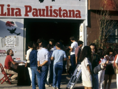 Foto da fachada do espaço cultural Lira Paulistana com uma multidão reunida na calçada. Pessoas conversam e folheiam livros, enquanto um homem sentado a uma mesa vermelha autografa obras. O letreiro “Lira Paulistana” está em destaque acima da entrada.