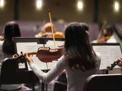 Foto de  músicos em uma apresentação ou ensaio orquestral, vistos por trás. O foco está em uma violinista de cabelos escuros, que segura seu violino e arco enquanto lê uma partitura apoiada em um suporte à sua frente. Ela veste uma blusa clara e está sentada em uma cadeira preta. Ao redor dela, outros músicos também tocam instrumentos de corda, como violinos e violas. Ao fundo, uma plateia desfocada, com assentos vazios.