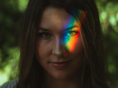Foto de uma mulher de cabelos escuros e olhos claros. Sobre um de seus olhos, a projeção de um arco-íris. Ao fundo, folhagens desfocadas. 