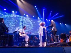 Foto de um palco durante um concerto. No centro, uma mulher negra, de cabelos crespos e loiros, está em pé diante de um microfone, usando um vestido escuro e um casaco branco e longo. Ao lado dela, um maestro de cabelos brancos usando um terno gesticula com uma das mãos erguidas, de frente para a orquestra. Atrás da cantora, um músico de roupas claras toca um violão sentado. À esquerda e à direita, membros da orquestra estão sentados com violinos, violoncelos e outros instrumentos.
