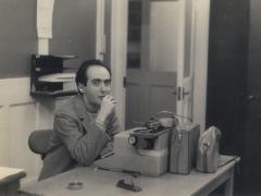 Foto em preto e branco de um homem, branco, de cabelos curtos, lisos e escuros, sentado em uma mesa com uma máquina de escrever à sua frente. Ele veste blazer e camiseta, tem os cotovelos apoiados sobre a mesa e uma das mãos próxima do rosto. Na mesa, há um cinzeiro, um grampeador e bolsas. Atrás dele, uma parede clara e uma porta aberta.