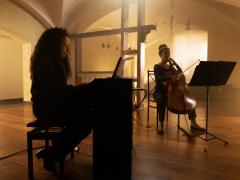 Foto de duas mulheres negras que tocam instrumentos em um ambiente iluminado por luz suave e amarelada. À esquerda, uma delas toca piano elétrico; à direita, a outra toca violoncelo, com uma partitura apoiada em um suporte à sua frente. As duas estão vestidas de preto e o ambiente tem piso de madeira e paredes claras.