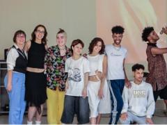 Foto de oito pessoas posando, de pé e sorrindo, em uma sala. Cinco das pessoas são brancas e três são rapazes negros, um deles está agachado e outro está de lado e segura um microfone em frente ao rosto. Todos usam roupas casuais como camisetas com alguma estampa e calças. O fundo tem paredes cinzas e uma projeção vermelha e amarela. 