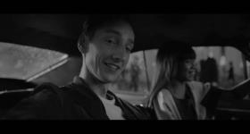 imagem em preto e branco de campanha publicitária em que aparecem um jovem branco, que sorri e olha para a camera, e ao fundo uma mulher, levemente desfocada, que também sorri enquanto olha a frente. eles estão dentro de um carro.