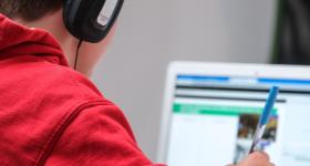 Foto de um garoto escrevendo, enquanto usa um laptop e fones de ouvido. O menino, que é branco e possui cabelo escuro, usa uma blusa de moletom vermelha e fones de ouvido pretos e cinzas. Ele segura com a mão direita uma caneta transparente de tampa azul. Ao fundo, há um laptop branco com a tela desfocada. Em último plano, está uma parede branca.