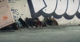 Foto de diversos pares de sapatos gastos encostados em uma parede. A parede apresenta alguns traços de grafite. A foto foi tirada na altura do chão.