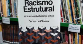 uma mão negra segura um livro, ao fundo, prateleiras com vários livros.