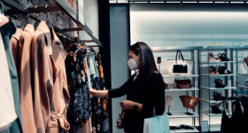 Foto de uma mulher escolhendo roupas em uma loja. A mulher é branca, possui cabelos lisos e escuros e está usando máscara. A roupa dela é preta e ela está com um relógio no braço esquerdo, no qual pendura uma sacola de compras. Essa mulher está escolhendo roupas entre várias opções penduradas em uma arara. Ao fundo, pode-se ver prateleiras com bolsas e sapatos.