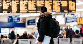 em um aeroporto, um homem está de pé, vestindo um caso preto e segurando um laptop na mão. Ele olha para um painel ao fundo, ligeiramente desfocado. à sua frente, também um pouco mais ao fundo, estão cadeiras brancas e pessoas sentadas de costas para ele.