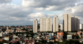 Foto de um bairro da região leste de Osasco. É possível observar uma comunidade composta por casas de baixo padrão cercada por prédios residenciais de classe média. O céu, ao fundo, tem muitas nuvens.