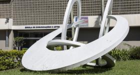 vista parcial da fachada do prédio da ECA; à frente a imagem de uma obra de arte, formada por quatro aneis entrelaçados em diferentes posições, na cor branca. Ao fundo, vista parcial dos cobogós que compõem parte da parede do prédio central.