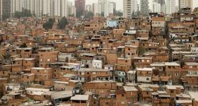Foto de uma favela tirada do alto. Diversas casas estão bem próximas e não estão rebocadas. Bem ao fundo, estão alguns prédios ca