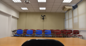 Foto de uma sala de aula. Em primeiro plano está uma mesa de madeira, de onde a foto foi tirada, e uma cadeira. Ao fundo, estão mais cadeiras, que são vermelha e azuis. A parede do fundo é bege e há um ventilador nela.