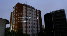 foto de um fim de tarde nublado, de um prédio branco e marrom. Nele, está projetada em luz a frase não há normalidade a que retornar.
