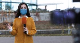 Foto de uma mulher, que é branca, tem cabelos lisos, longos e escuros e veste laranja. segurando um microfone e usando máscara. Desfocada, à direita da imagem, tem-se uma câmera direcionada à mulher.