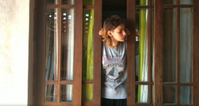 mulher jovem, branca, de cabelos lisos, castanhos e na altura do ombro, abra com as duas mãos uma porta de madeira e vidro. Ela veste uma camiseta cinza, estampada e olha para o lado direito.