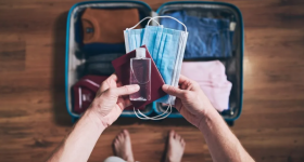 ao centro da imagem, duas mãos seguram duas máscaras, um frasco de alcool em gel e um passaporte. Ao fundo, uma mala aberta com roupas dobradas