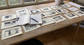 Foto de bancada de madeira sobre a qual há uma série de desenhos feitos em nanquim sobre papel branco. Eles estão organizados em linhas e colunas, como se indicassem uma sequência de cenas. Sobre alguns desenhos, no lado esquerdo da bancada, há um caderno com desenhos ainda inacabados de um storyboard, junto com uma lapiseira e uma borracha. 