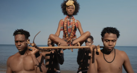 Foto mostra uma mulher jovem e negra de cabelos escuros e crespos em um corte redondo, como uma coroa. Ela usa um adorno de tecido no cabelo, lembrando uma tiara com flor, e roupas com grafismos geométricos em tons de azul, vermelho e amarelo. Ela está sentada em posição de lótus sobre uma espécie de maca, que é carregada nos ombros por quatro homens negros e jovens. Ao fundo, o mar e o céu, como se as pessoas estivessem saindo da água.