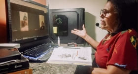 foto de uma senhora 0 branca, de cabelos de tamanho médio e levemente ondulados, oculos, que veste uma blusa vermelha. Ela olha para um monitor de computador, em que conversar via chamada de vídeo com outras duas pessoas. Ela está sentada em uma escrivaninha e, à frente dela, há um livro aberto.