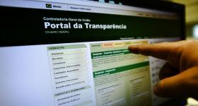 Foto da tela de um computador. Na tela, o site do Portal da Transparência do governo federal. O site possui colunas em cores verde claro e escuro. No canto direito da imagem aparece uma mão com o dedo indicador apontando para a tela. 