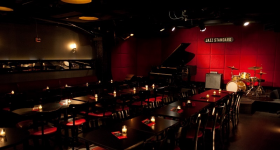 Foto de uma sala de jazz vazia, com várias mesas juntas e ornadas com velas acesas. Ao fundo, sobre um pequeno palco e de frente para uma parede vermelha, encontram-se um piano, uma caixa de som, um piano e uma bateria.
