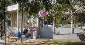 Foto da fachada do prédio do CMU, que tem cores claras. Na entrada, há uma escada e uma rampa. Na frente do prédio, além de muitas árvores, há alguns bancos, bicicletas e telefones “orelhões”. A placa do prédio tem um tom rosa forte e a sigla CMU em letras brancas.