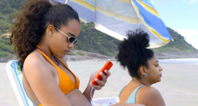 Imagem do filme "Receita de Caranguejo". Há, na foto, duas mulheres pretas em uma praia. A mulher em primeiro plano, sentada e usando óculos e biquíni amarelo, olha para seu celular de capa laranja. A outra, de biquíni e brinco azul, olha para a paisagem. Ao fundo, pode-se observar uma montanha e parte do mar.